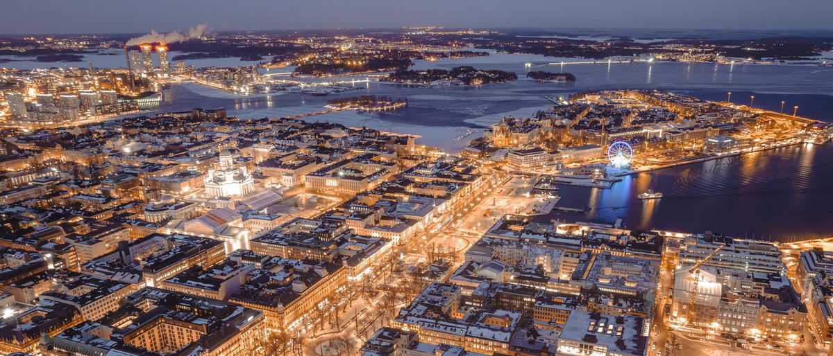 Luftbild der Innenstadt von Helsinki an einem Winterabend, beleuchtete Straßenzüge und vereistes Hafenwasser